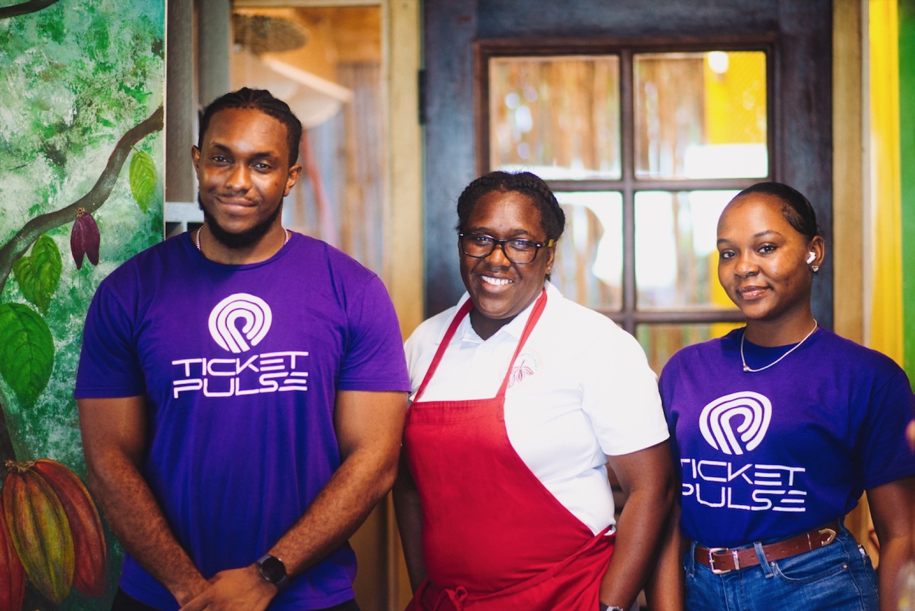 Two Pulse crew in TicketPulse shirts with the owner of a cocoa shop, in front of a painted cocoa mural.